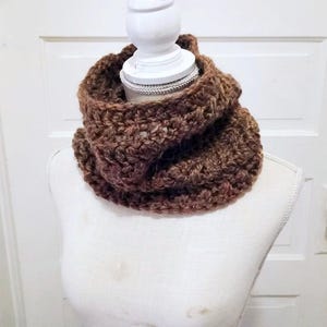 May include: A chunky, hand-crocheted infinity scarf in a variegated brown yarn. The scarf features a thick, textured stitch pattern and is draped around the neck of a white mannequin. The yarn displays various shades of brown, from light to dark.