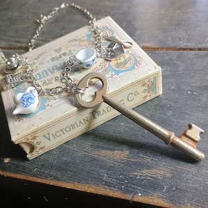 Può includere: Una collana a catena in metallo color argento con diversi ciondoli in miniatura, tra cui una teiera e una tazzina in ceramica bianca con motivi floreali blu, un cucchiaino in metallo e una grande chiave antica fissata alla catena.
