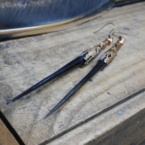May include: A pair of long, black, pointed earrings with gold accents. The earrings are made of wood and have a delicate, intricate design.