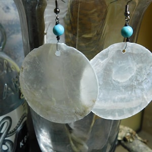 May include: A pair of earrings with white shell pendants and turquoise beads. The earrings are hanging from silver hooks.