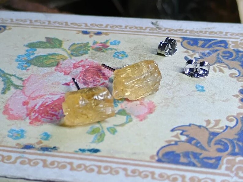 Puede incluir: Un par de pendientes de bot&oacute;n de citrino en bruto con postes y cierres de plata. Los pendientes son de color amarillo claro y tienen un aspecto rugoso y texturizado. Se exhiben sobre una superficie con estampado floral.