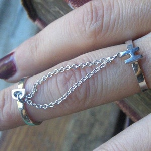 May include: A silver ring with a chain attached to a charm with the letter "H" on it. The ring is worn on a finger with dark purple nail polish.