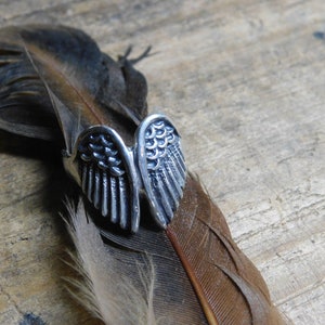 May include: A silver ring with a detailed design of two angel wings. The ring is resting on a brown feather.