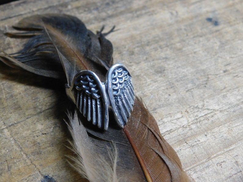 May include: A silver ring with a detailed design of two angel wings. The ring is resting on a brown feather.