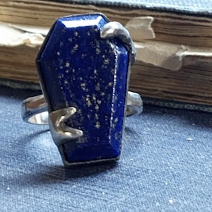 May include: A silver ring featuring a blue lapis lazuli stone shaped like a coffin. The stone has a sparkly, glittery texture.