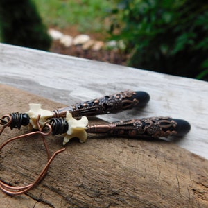May include: A pair of dangle earrings with a black and copper filigree design. The earrings feature a white bone-like charm and a black bead.