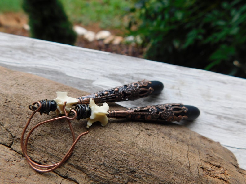 May include: A pair of dangle earrings with a black and copper filigree design. The earrings feature a white bone-like charm and a black bead.