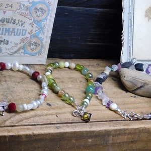 May include: Three beaded bracelets with different colored stones and charms. The bracelets are on a wooden surface. The bracelet on the left has red, white, and brown beads with a silver piano charm. The middle bracelet has green and white beads with a silver charm. The bracelet on the right has a variety of colored stones and a silver clasp.