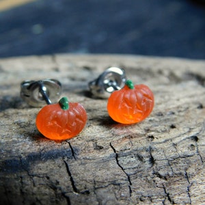 May include: A pair of silver stud earrings with orange pumpkin charms. The pumpkins have green stems and carved faces.