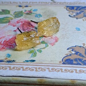 Puede incluir: Un par de pendientes de bot&oacute;n de citrino en bruto. Los pendientes son de color &aacute;mbar claro y tienen una textura rugosa y natural. Los postes son oscuros. Los pendientes se exhiben sobre una superficie con estampado floral con flores rosas y azules.