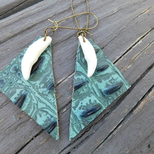 May include: A pair of earrings with a teal leather triangle shape and a white tooth charm. The earrings have a bronze wire hook.