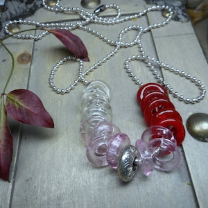 May include: A silver chain necklace with a pink and red glass bead charm. The charm is made up of several round beads that are stacked on top of each other. The beads are translucent and have a slight shimmer to them.