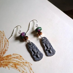 May include: A pair of handmade earrings with silver hooks. Each earring features a dark gray, textured rectangular pendant with a cross design. The earrings also include ruby and green beads, and copper-colored beads.