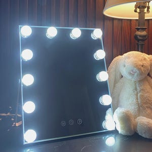 May include: A rectangular vanity mirror with twelve integrated LED bulbs arranged along the perimeter. The mirror features a touch-sensitive control panel at the bottom center with three circular icons labeled M, a power symbol, and P.