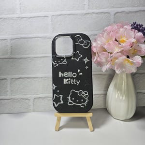May include: Black phone case featuring the Hello Kitty character, stars, and bows. The case has a clear camera opening and is displayed on a small wooden easel. The words "hello Kitty" are in white lettering.