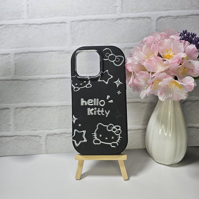 May include: Black phone case featuring the Hello Kitty character, stars, and bows. The case has a clear camera opening and is displayed on a small wooden easel. The words "hello Kitty" are in white lettering.