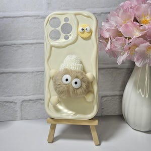 May include: Cream-colored phone case with a textured surface, featuring a fluffy, light brown creature with large googly eyes and a small white knitted hat. Additional yellow and white embellishments add to the whimsical design. The case is displayed on a small wooden easel.