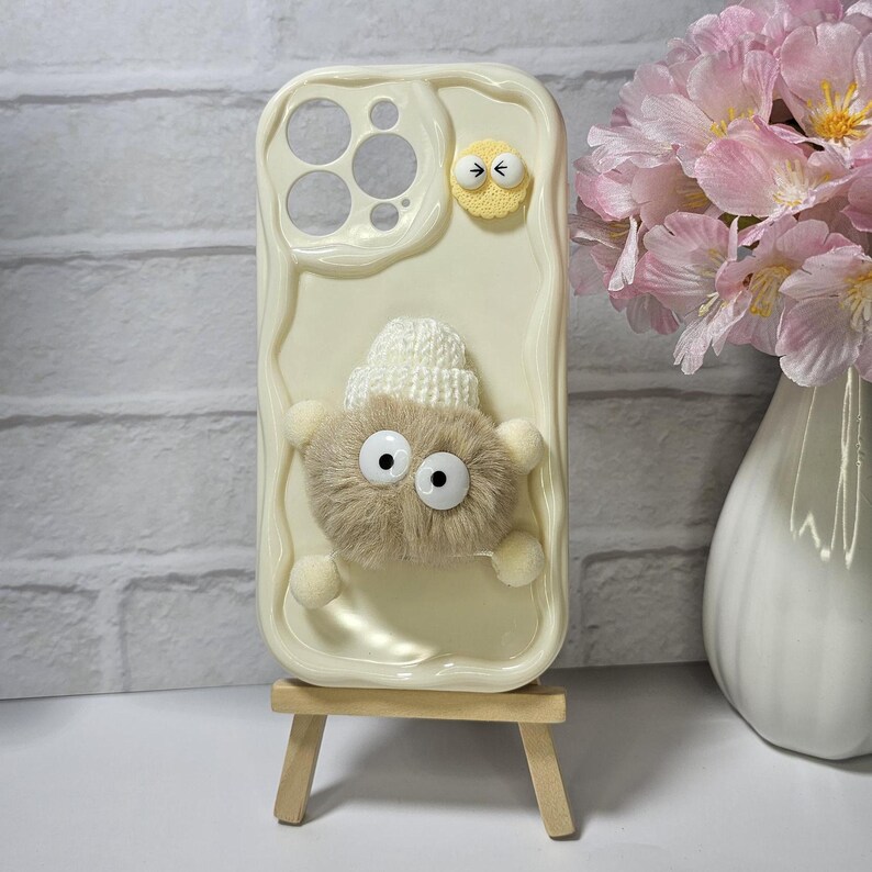 May include: Cream-colored phone case with a textured surface, featuring a fluffy, light brown creature with large googly eyes and a small white knitted hat. Additional yellow and white embellishments add to the whimsical design. The case is displayed on a small wooden easel.