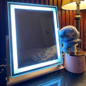Puede incluir: Un espejo de tocador cuadrado con un borde brillante e iluminado. El espejo está sobre un soporte y tiene una base rosa claro. Un pequeño muñeco de peluche con pelaje azul y una bufanda azul está sobre una vela rosa.