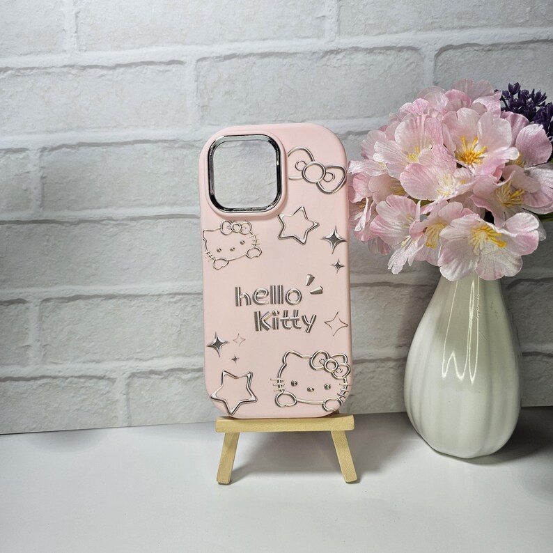May include: A light pink phone case featuring Hello Kitty designs and the text "hello kitty". The case has a silver camera cutout and silver accents of stars and bows. The case is displayed on a small wooden easel.