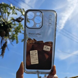 May include: Clear phone case with a black border, featuring a graphic of iced black coffee with ice cubes. The case has text that reads "Black Coffee" and "Daily coffee moments". Additional text includes "Milk", "Soda", and "Sugar".