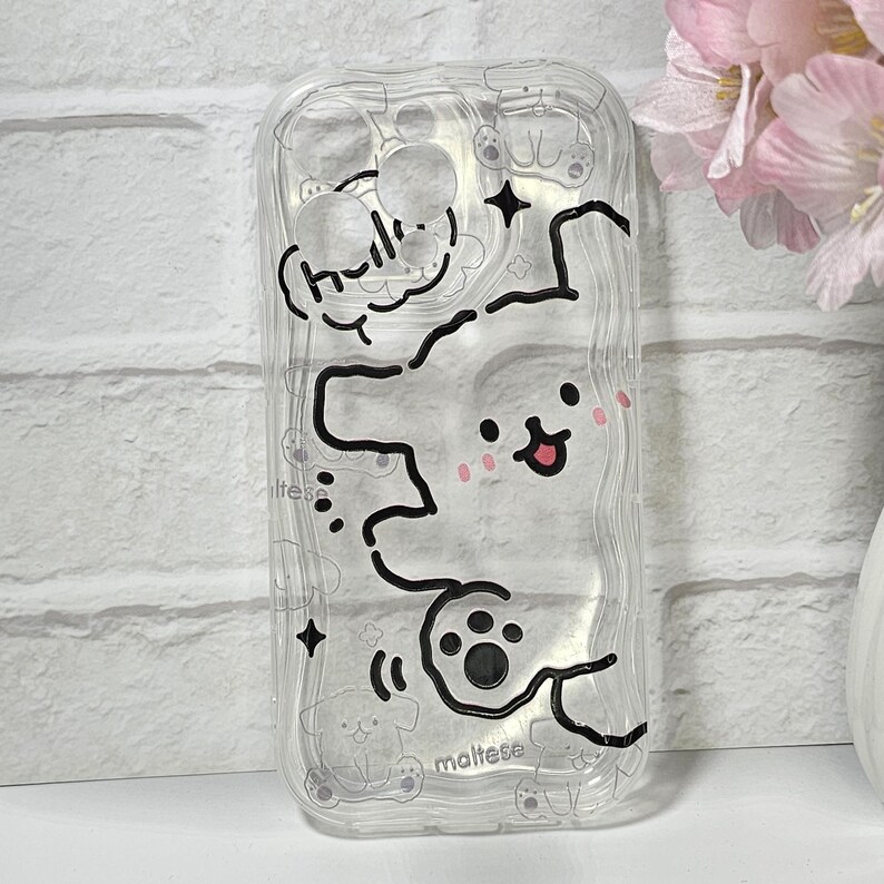 May include: Clear phone case with a cartoon Maltese dog design. The case features a white outline of a dog with pink cheeks, paw prints, and the word "hello" in a speech bubble. The word "maltese" is also visible.