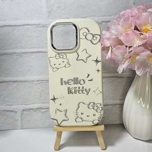 May include: Off-white phone case featuring Hello Kitty designs and the text "hello Kitty". The case has a camera cutout and is decorated with stars and bows. The case is on a small wooden easel.