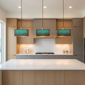 Puede incluir: Tres lámparas colgantes rectangulares con pantallas turquesas y doradas cuelgan sobre una isla de cocina. La cocina cuenta con gabinetes de madera clara, una encimera blanca y una estufa de acero inoxidable. Las luces añaden un toque moderno.