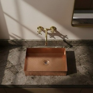 Peut inclure: Un évier rectangulaire en cuivre avec une texture martelée, posé sur un comptoir sombre. Un robinet doré avec des poignées en croix est monté au-dessus de l'évier. L'évier a un drain au centre.