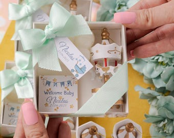 May include: White gift boxes tied with mint green ribbons, each containing a carousel figurine and a small card that says "Welcome Baby Nicolas 25.06.2023". Additional carousel figurines are visible.