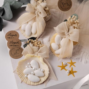 May include: Seashell favours filled with white sugared almonds and pearls, decorated with beige bows and dried flowers. Includes small round labels with text. Yellow starfish accents the display.