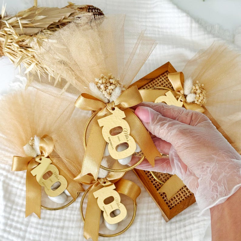 May include: Gold-toned party favors featuring a circular frame, a satin ribbon bow, and a cutout number 100 with the text Mila's Birthday. Each favor is wrapped in gold tulle fabric and decorated with small dried white flowers and grasses.