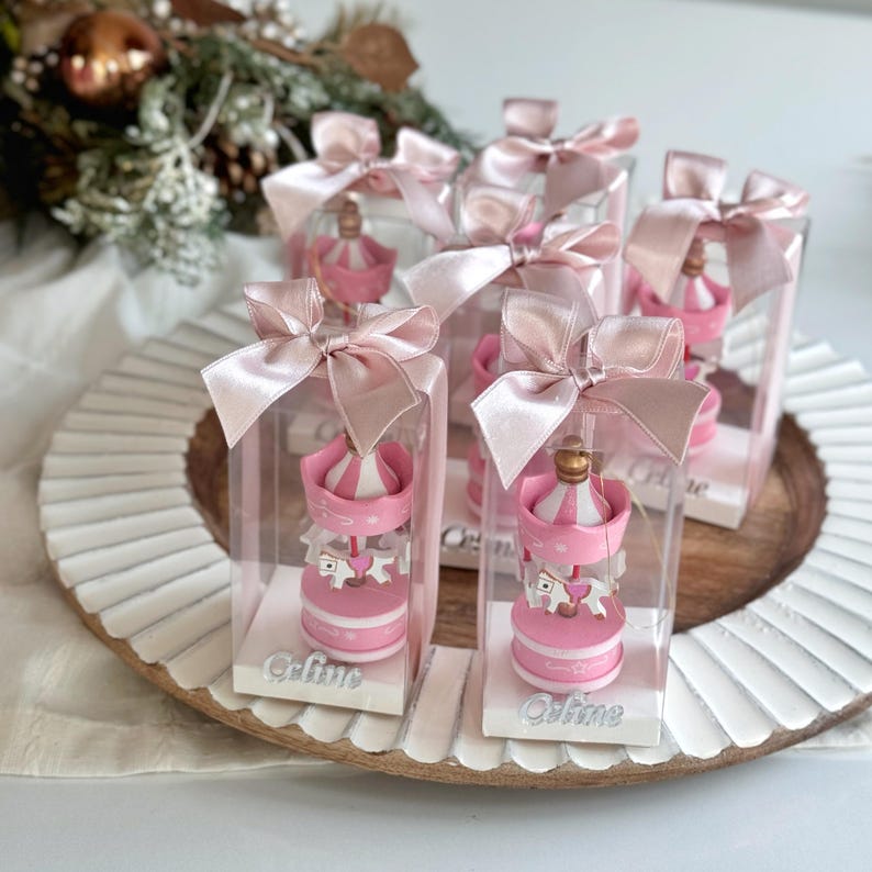 Pink carousel party favors in clear gift boxes with satin ribbons and personalized acrylic names – ideal for baby showers or baptisms. Close-up of pink carousel keepsakes with gold and silver bows, featuring custom names and elegant acrylic details. Colorful carousel favors (red, blue, green, yellow) with festive ribbons, designed as personalized welcome baby or birthday gifts.