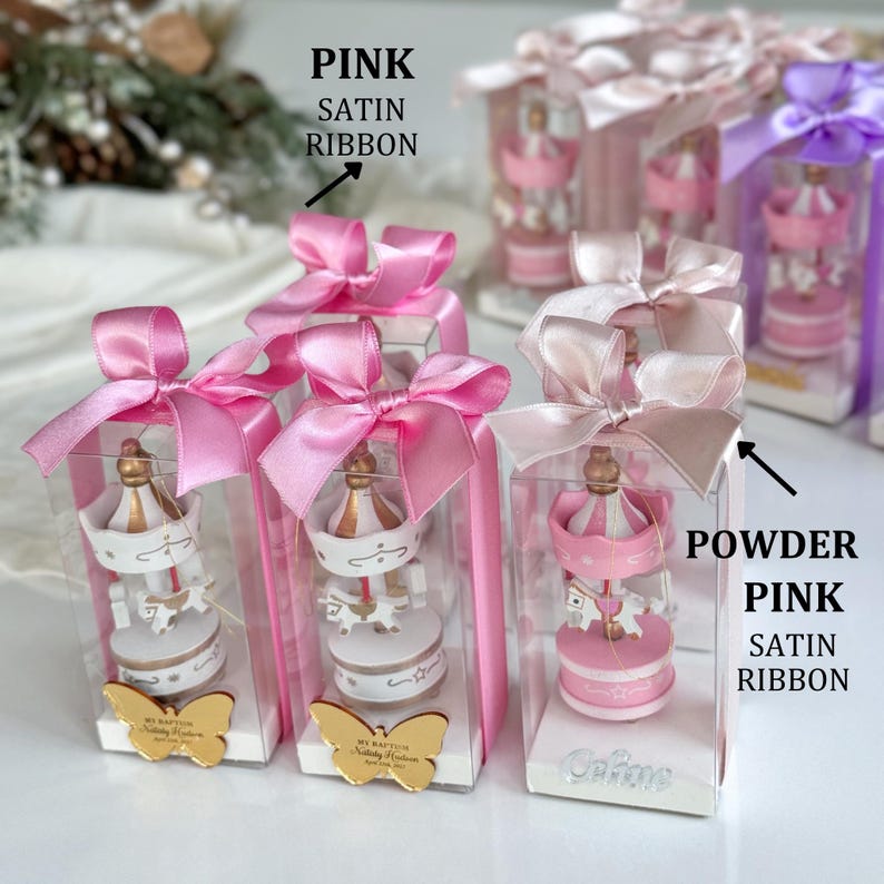 Pink carousel party favors in clear gift boxes with satin ribbons and personalized acrylic names – ideal for baby showers or baptisms. Close-up of pink carousel keepsakes with gold and silver bows, featuring custom names and elegant acrylic details. Colorful carousel favors (red, blue, green, yellow) with festive ribbons, designed as personalized welcome baby or birthday gifts.