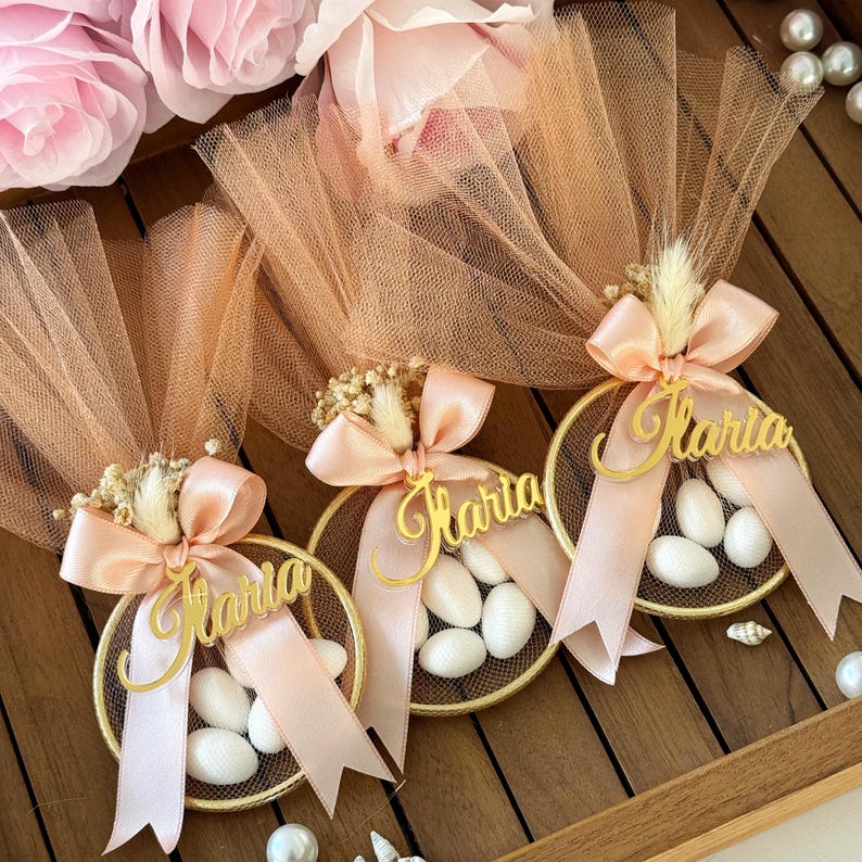 Personalized gold baptism favor with soft peach tulle, gold acrylic name tag ‘Ilaria’, satin ribbon bow, and sugared almonds – elegant handmade bomboniere for girl baptism or baby shower