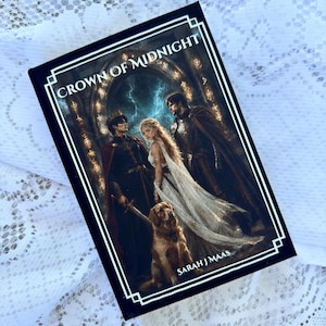 Pode incluir: Um livro de capa dura intitulado Crown of Midnight de Sarah J. Maas. A capa apresenta uma ilustração de duas figuras em trajes escuros e uma mulher com um vestido branco diante de um arco de pedra, com um cão dourado em primeiro plano.