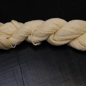 Puede incluir: Un ovillo de hilo blanquecino, bien torcido y enrollado, descansa sobre una superficie oscura. El hilo parece suave y natural, adecuado para proyectos de tejido o ganchillo. El color neutro sugiere versatilidad para diversos diseños artesanales.