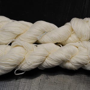 Puede incluir: Dos madejas de hilo blanco roto se presentan sobre una superficie oscura. El hilo está bien enrollado, mostrando un aspecto suave y texturizado. Las madejas están colocadas una al lado de la otra, lo que sugiere que forman parte de un conjunto o están destinadas a un proyecto más grande. Ideal para tejer o hacer ganchillo.