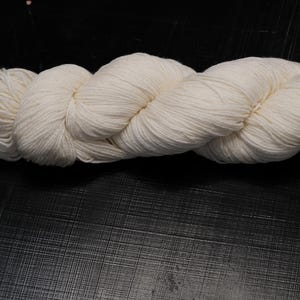 Puede incluir: Un ovillo de lana de color crema, bien enrollado y listo para tejer o hacer ganchillo. La lana es de un color suave y neutro, perfecta para varios proyectos. La textura parece suave y consistente, ideal para crear prendas o accesorios.