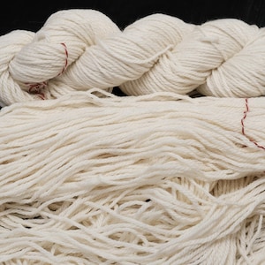 Puede incluir: Dos madejas de hilo blanco roto. Una madeja está bien enrollada, mientras que la otra está dispuesta sueltamente. Ambas están atadas con un hilo rojo fino. El hilo parece estar hecho de una fibra natural, adecuado para proyectos de tejido o ganchillo.