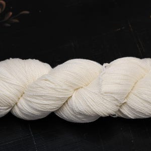 Puede incluir: Un ovillo de hilo blanco roto, bien enrollado y retorcido, sobre una superficie oscura. El hilo parece suave y texturizado, adecuado para proyectos de tejido o ganchillo. El color neutro sugiere versatilidad para diversos proyectos.
