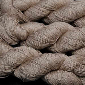 Puede incluir: Primer plano de varios ovillos de hilo beige. El hilo está enrollado firmemente en formas suaves y redondeadas, con hebras visibles y un aspecto ligeramente texturizado. El color es un tono cálido y neutro, adecuado para varios proyectos de tejido o ganchillo.
