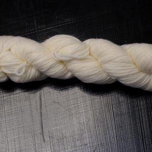 Puede incluir: Un ovillo de lana de color crema, fuertemente retorcido en forma de cuerda. La lana es de color neutro y parece suave y esponjosa. Una pequeña etiqueta rosa está adherida a un extremo. Ideal para tejer o hacer ganchillo.