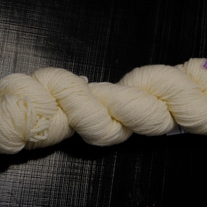 Puede incluir: Un ovillo de lana de color crema, bien enrollado y retorcido, descansa sobre una superficie oscura. La lana parece suave y esponjosa, con unas pequeñas etiquetas moradas visibles. Adecuado para proyectos de punto o ganchillo.
