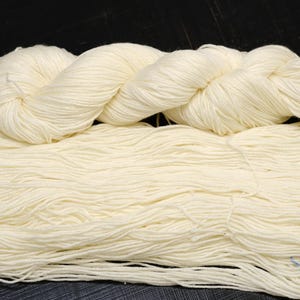 Puede incluir: Un ovillo de lana de color crema, con una parte superior retorcida y una sección inferior suelta y fluida. La lana parece suave y es adecuada para proyectos de tejido o ganchillo. El color es un tono crema natural.