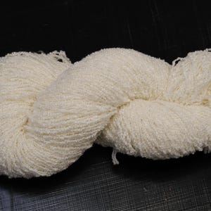 Puede incluir: Un ovillo de lana de color crema, bien enrollado y texturizado, sobre una superficie oscura. La lana parece suave y esponjosa, ideal para proyectos de punto o ganchillo. La imagen resalta el color y la textura naturales de la fibra.