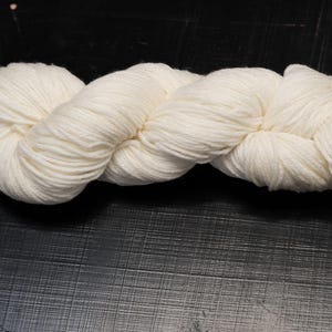 Puede incluir: Un ovillo de lana de color crema, bien torcido y enrollado, descansa sobre una superficie oscura. La lana parece suave y esponjosa, ideal para proyectos de tejido o ganchillo. El color neutro es adecuado para varios proyectos.