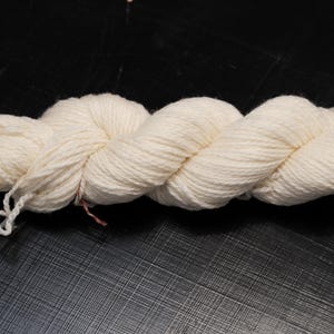 Puede incluir: Un ovillo de hilo blanco roto, bien torcido y listo para tejer o hacer ganchillo. El hilo es de color natural, con un aspecto ligeramente texturizado. El ovillo descansa sobre una superficie oscura y texturizada.