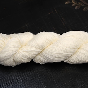 Puede incluir: Un ovillo de hilo blanquecino, bien enrollado y listo para tejer. El hilo es de color crema, con hebras visibles y una textura suave. El ovillo descansa sobre una superficie oscura, resaltando su color y forma.