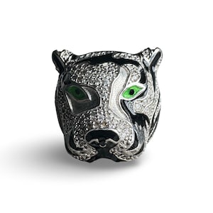 May include: A silver ring with a black and white tiger head design. The tiger has green eyes and is set with small, clear stones.